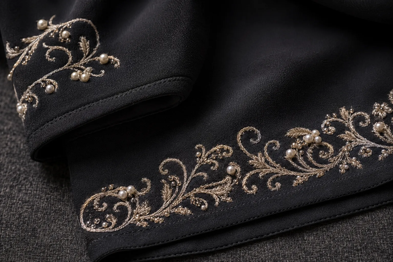 Nahaufnahme einer schwarzen Abaya: goldfarbene Ranken- und Blütenstickerei mit eingearbeiteten Perlen entlang von Ärmelaufschlag und Saum