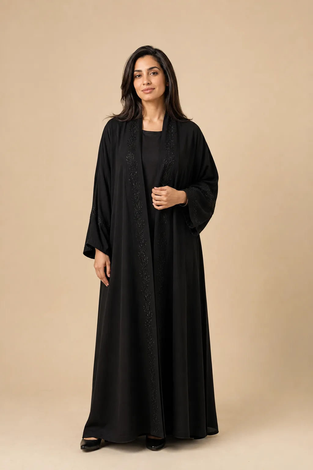 Frau in einer offenen schwarzen Abaya mit floraler Stickerei an Saum und Ärmeln, darunter ein schwarzes Kleid; Haare und Gesicht sind unbedeckt