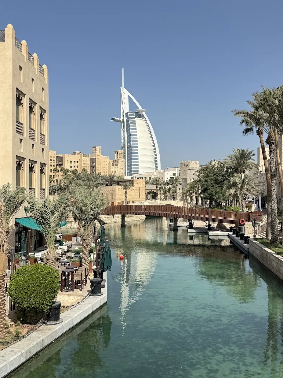 Blick über den Kanal von Madinat Jumeirah: links ein Gebäude im arabischen Stil mit Windturm und eine Terrasse unter Palmen, in der Mitte eine hölzerne Fußgängerbrücke über dem türkisfarbenen Wasser, dahinter das segelförmige Burj Al Arab vor wolkenlosem Himmel