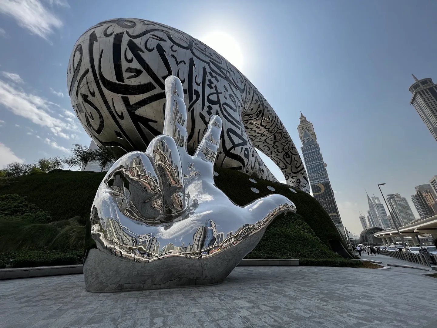 Das ringförmige Gebäude des Museum of the Future mit arabischer Kalligrafie auf der Fassade, davor die spiegelnde Chrom-Skulptur einer geöffneten Hand; rechts dahinter die Hochhäuser an der Sheikh Zayed Road