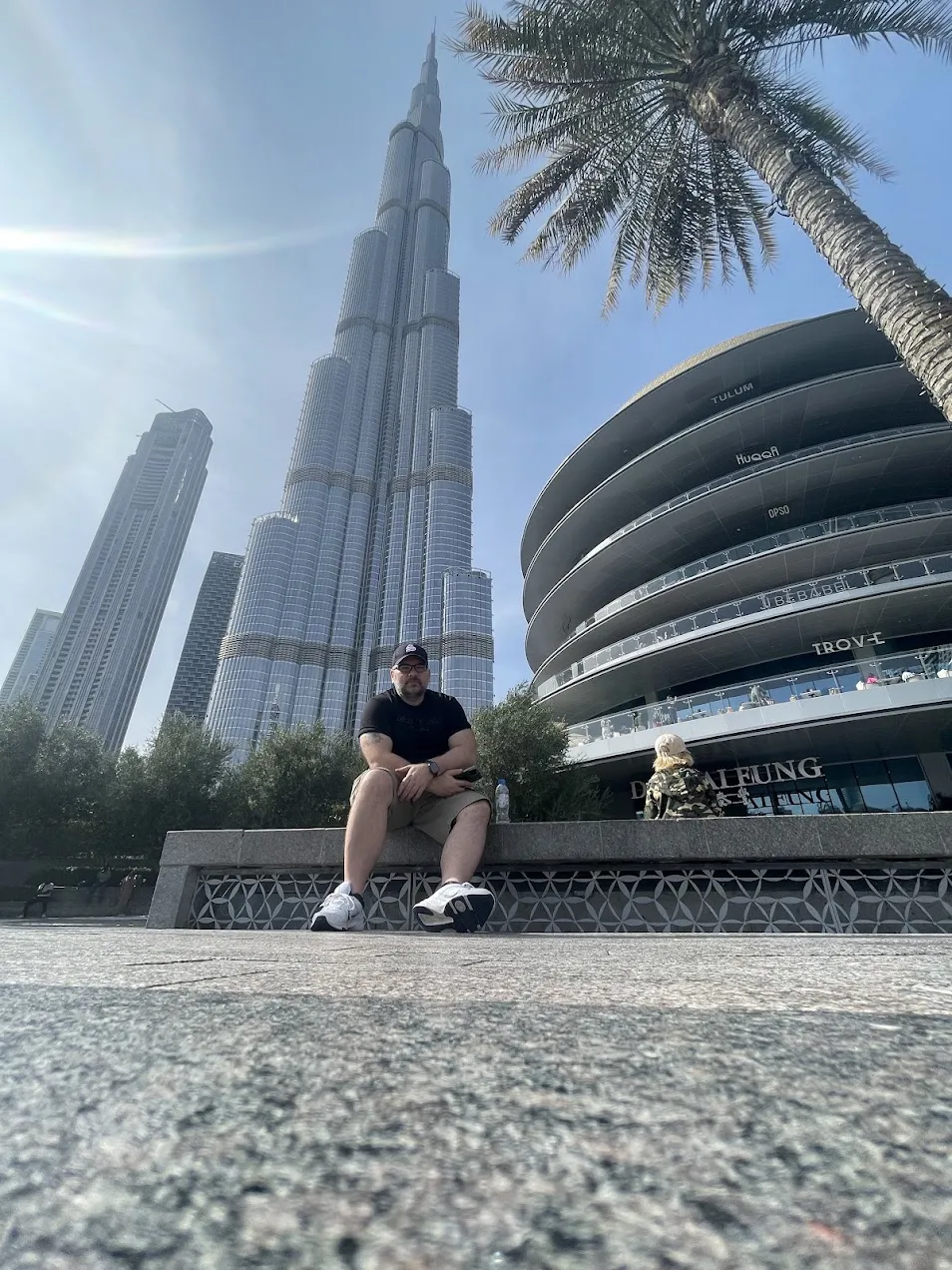 Sandro sitzt auf einer niedrigen Mauer vor dem Burj Khalifa, links der Turm in voller Höhe, rechts eine Palme und die geschwungene Fassade der Fashion Avenue