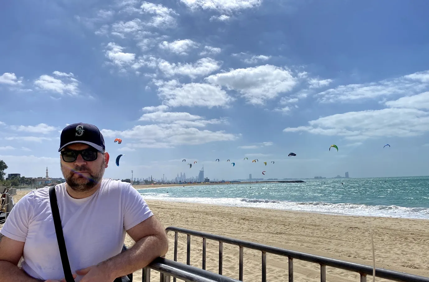 Sandro mit Kappe und Sonnenbrille an der Strandpromenade am Kite Beach, dahinter Dutzende Kitesurf-Schirme über dem Meer und in der Ferne die Skyline mit dem Burj Al Arab